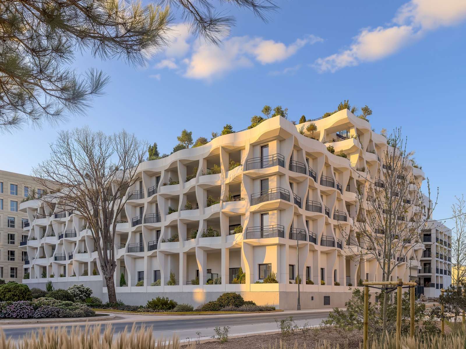 Exterior view of the Secret Gardens residential building in Montpellier featuring white organic undulating facades and lush rooftop gardens at sunset.