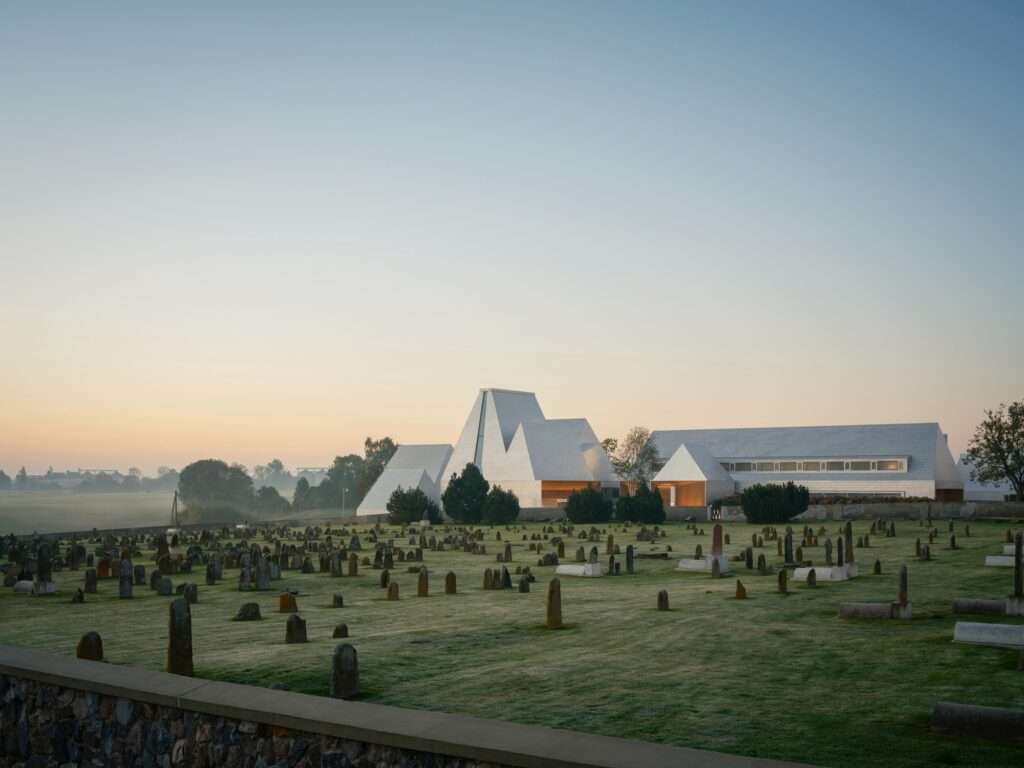 A wide exterior shot of The Lost Shtetl Museum in Šeduva at sunset, featuring the shingle-style aluminum facade overlooking the historic Jewish cemetery.