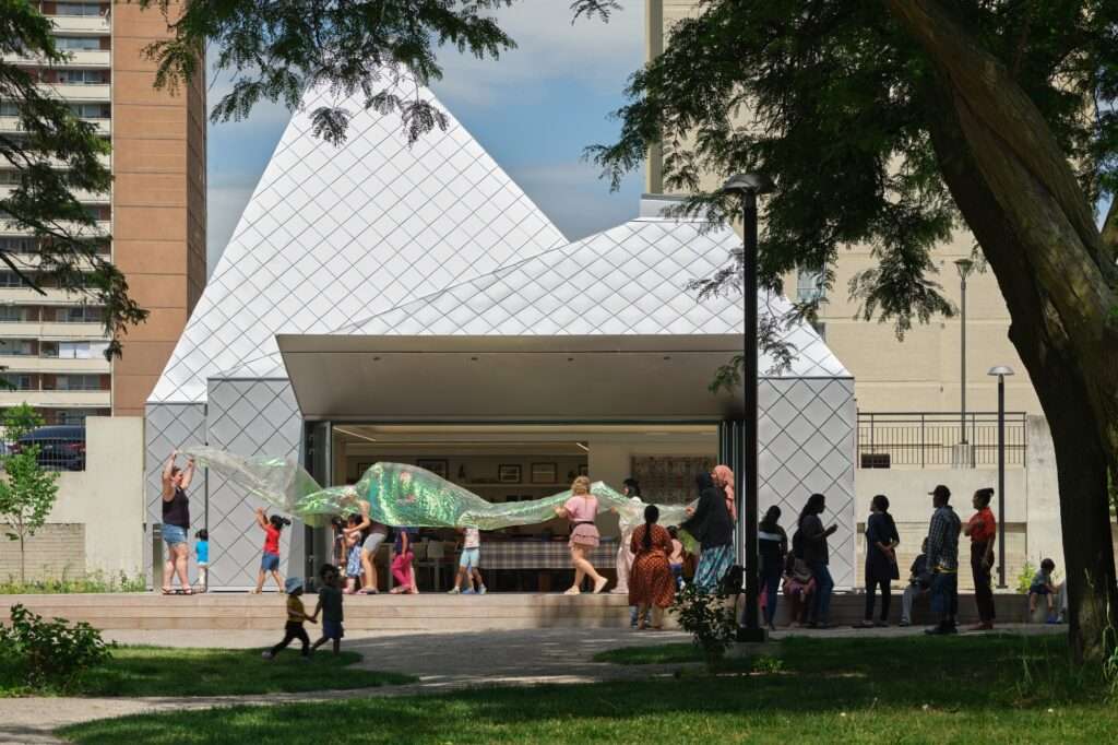 Children playing with a shimmering green fabric on the open deck of the diamond-patterned aluminum pavilion at Mabelle Park.