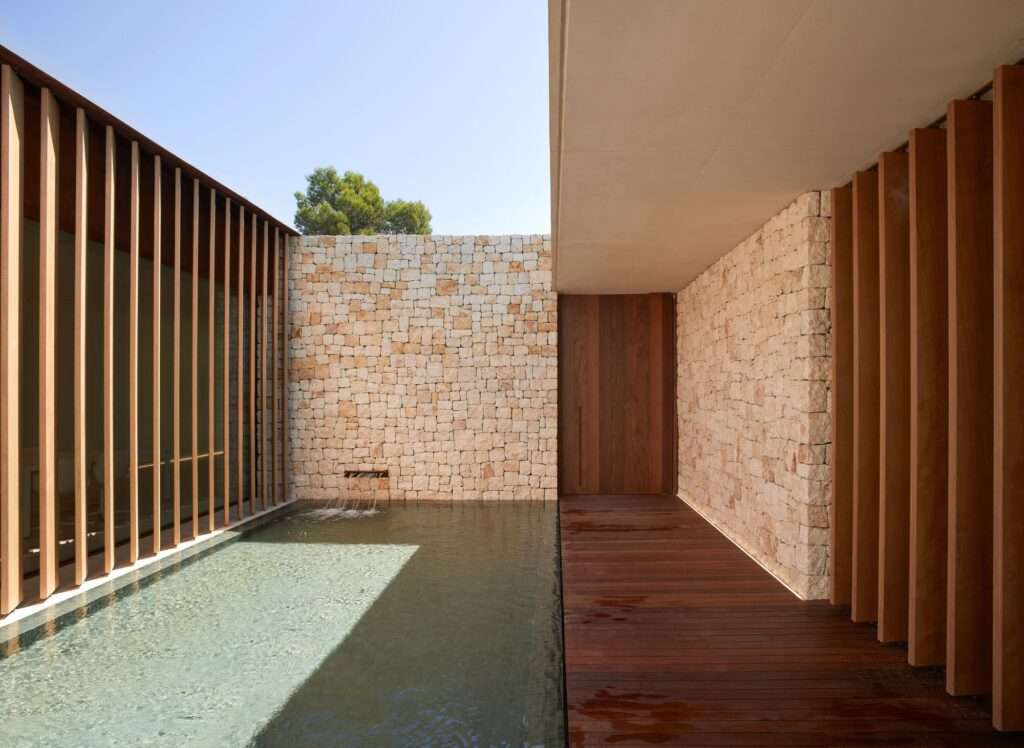Interior courtyard featuring a natural stone wall, a shallow water pool, and vertical wooden slats under a clear sky.