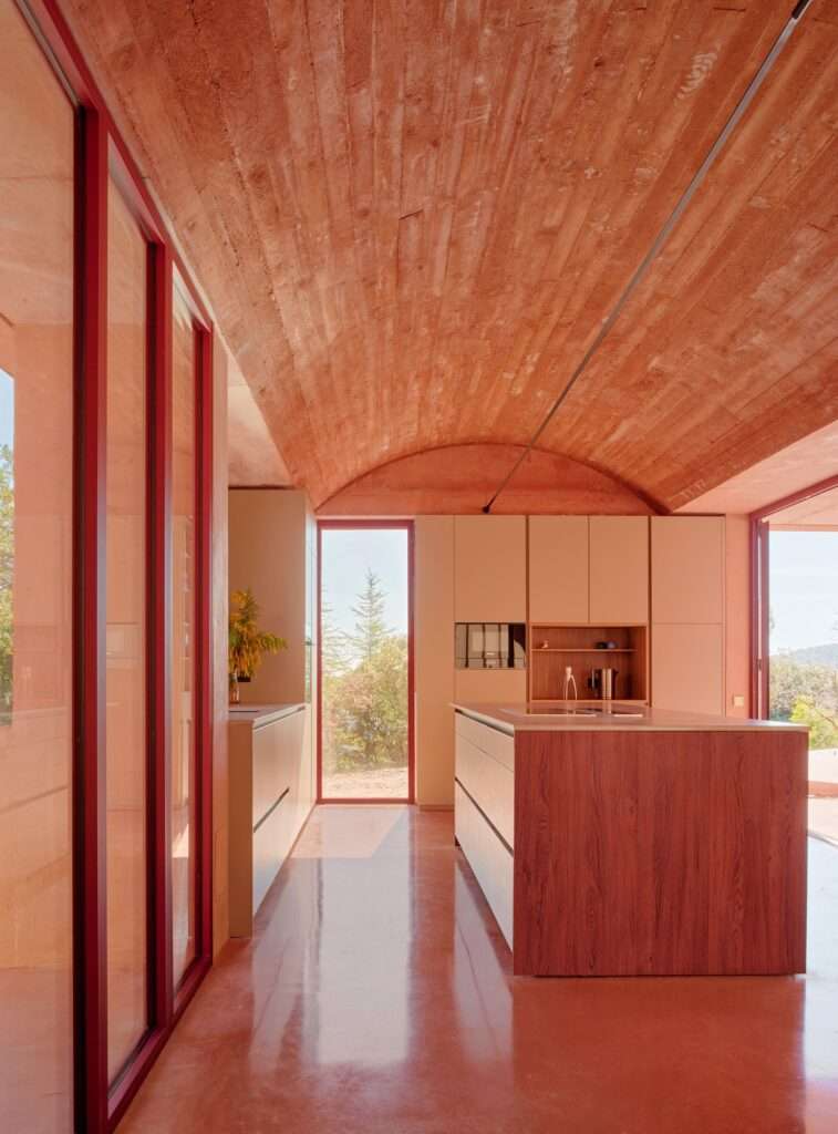 Red-tinted concrete kitchen interior at Casa Sota la Mola with a Catalan vault ceiling, timber-textured surfaces, a central wooden island, and floor-to-ceiling glass windows.