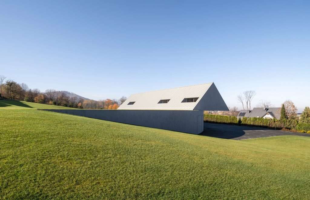 Side view of a modern minimalist gabled house embedded into a steep green grass slope under a clear blue sky.