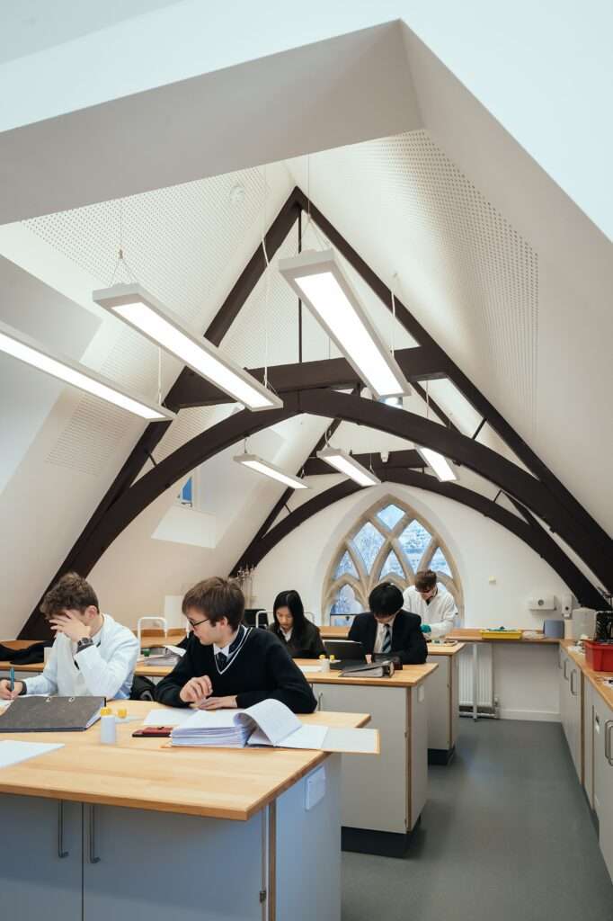 Students in lab coats working inside an attic physics laboratory with white walls, exposed dark structural timber arches, and an ornate stone Gothic window at the far end.