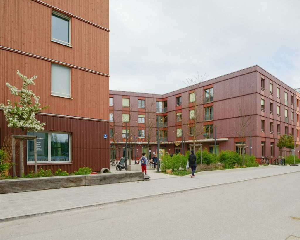 Neighborhood square and mixed-use residential buildings with a ground-floor cafe and reddish wood facade in Freiham, Munich.