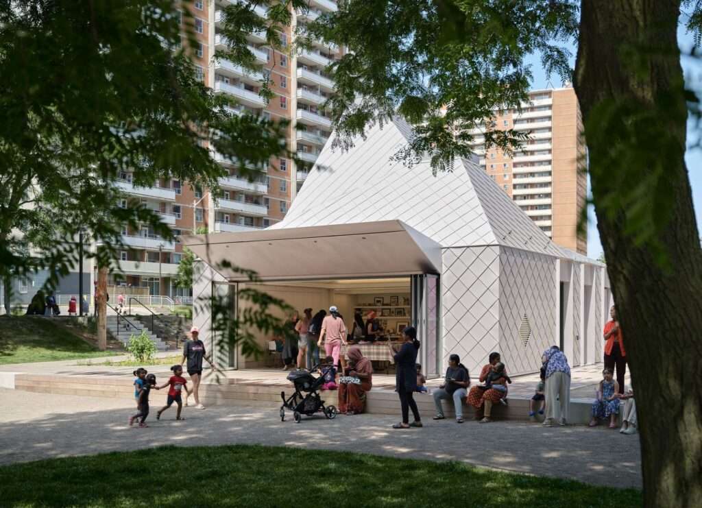 Neighbors socializing on the wide wooden deck of the modern metallic pavilion with residential tower blocks visible in the background.