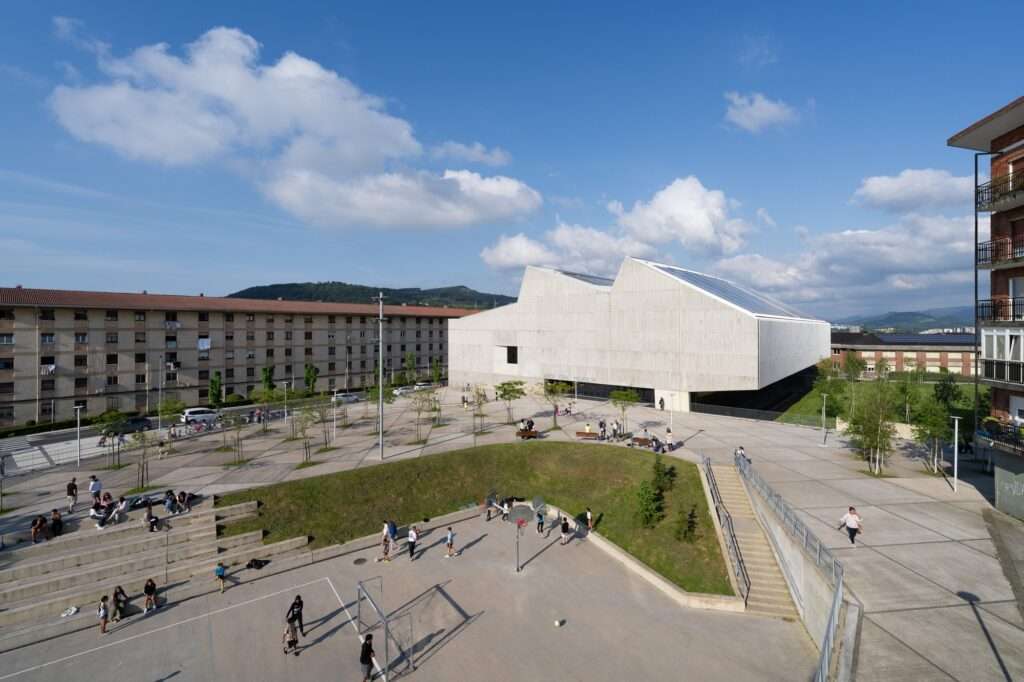 Wide shot of an outdoor sunken basketball and football court with concrete tiered spectator seating, framed by the modern sports complex building and residential apartments.