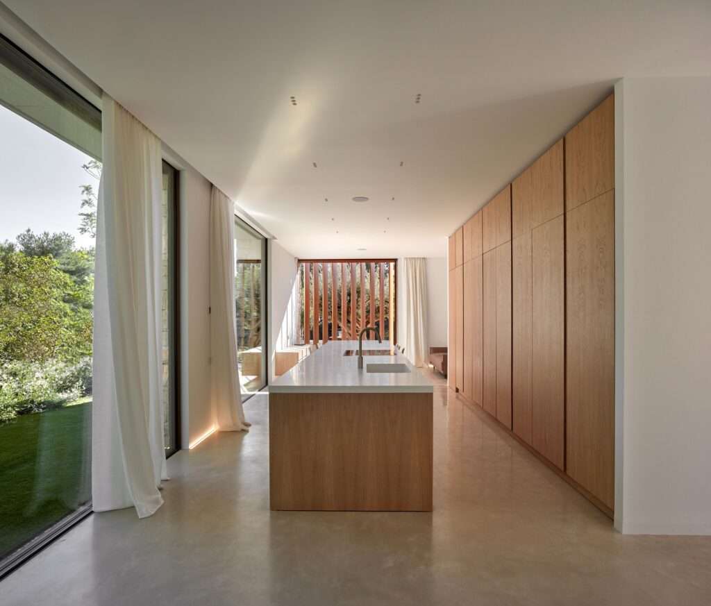 Minimalist kitchen interior with a long wooden island, polished concrete floors, and large windows looking out to the garden.