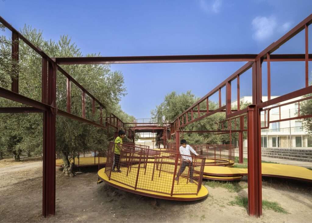 Ground-level view of children playing on a spinning yellow carousel under a dark red steel grid framework.