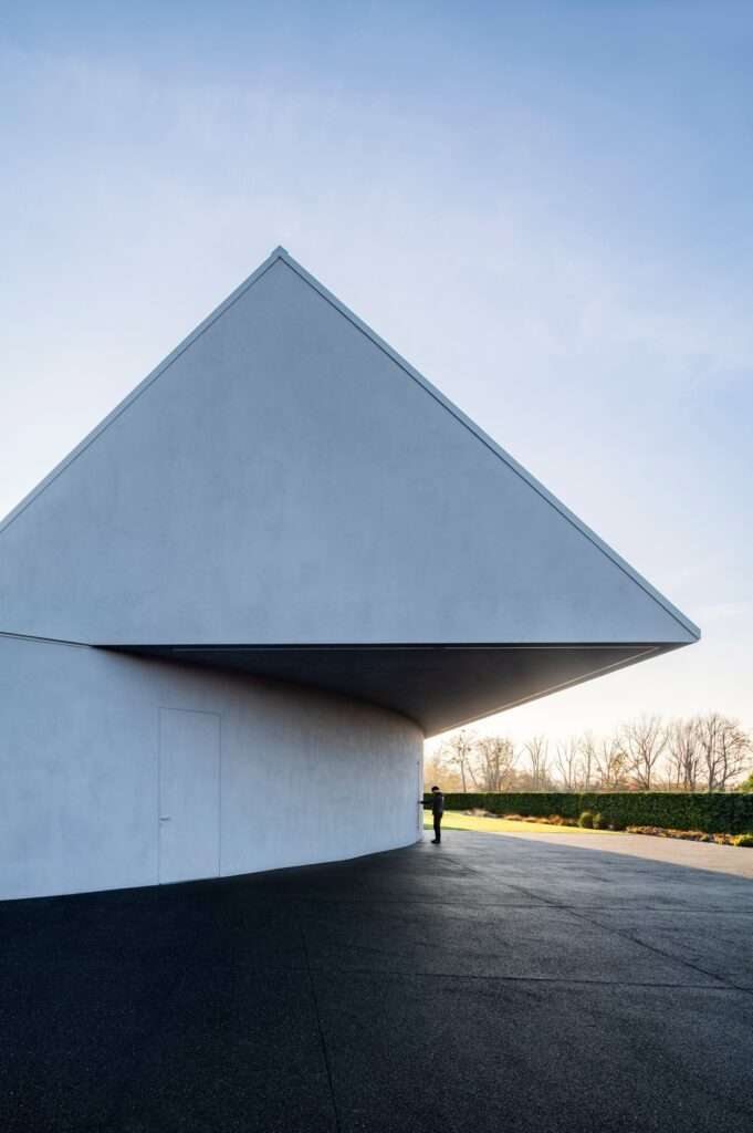 Close-up architectural shot of the sharp cantilevered gabled roof and curved concrete wall of the Yaw House, with a person standing for scale.