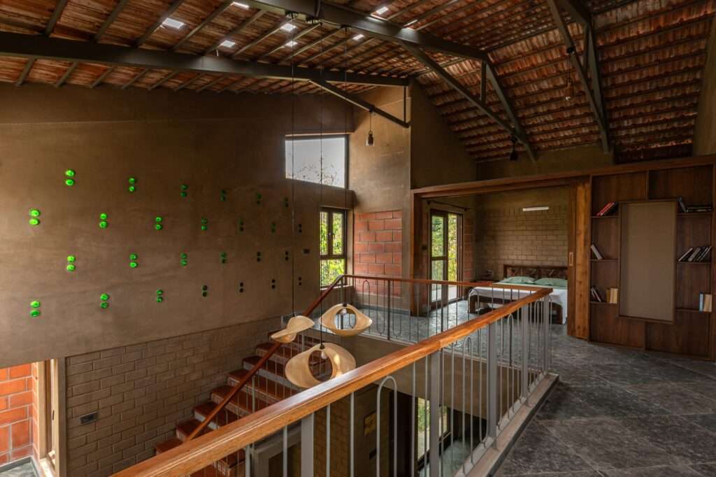 Upper level interior balcony corridor in Svatamman project overlooking a double height space, showcasing a bedroom with wooden sliding doors, custom pendant lights, and a feature wall with embedded green glass bottles.