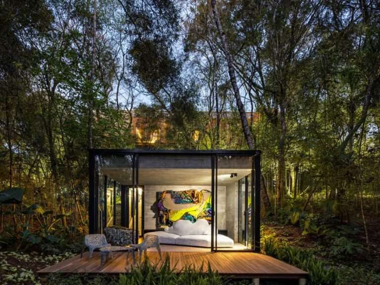 Evening view of House 17-JB by Luis Volpato in Curitiba, featuring a glass pavilion with a colorful art piece integrated into a dense forest.