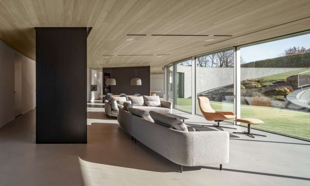 Minimalist interior design of the open-plan living room with curved wooden ceilings, large floor-to-ceiling glass windows, and views of a landscaped slope.