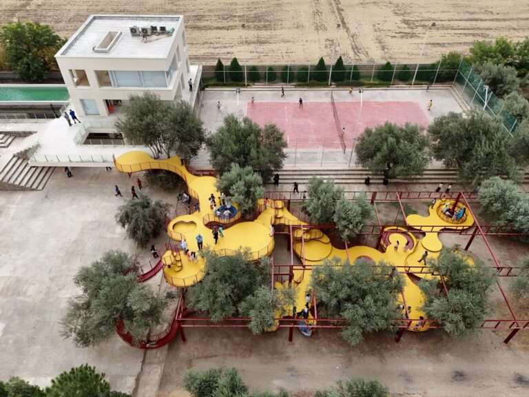 Aerial drone view of a yellow elevated playground built within an olive grove next to a modern building and sports courts in Minodasht.