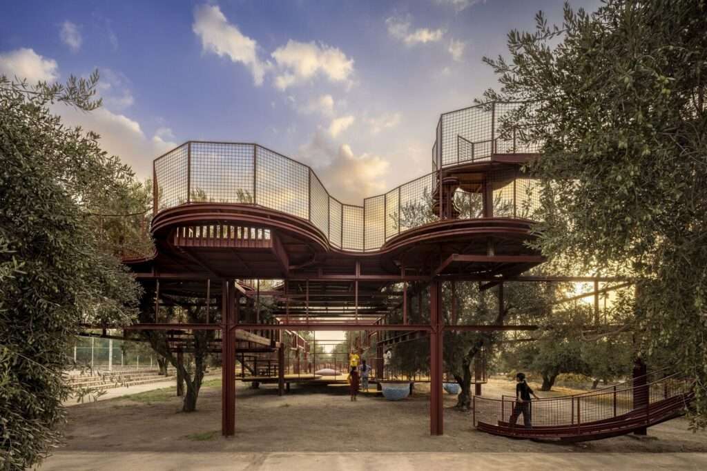Warm sunset view of the playground structure showing the underlying red steel pillars, mesh railings, and upper platforms in an olive orchard.
