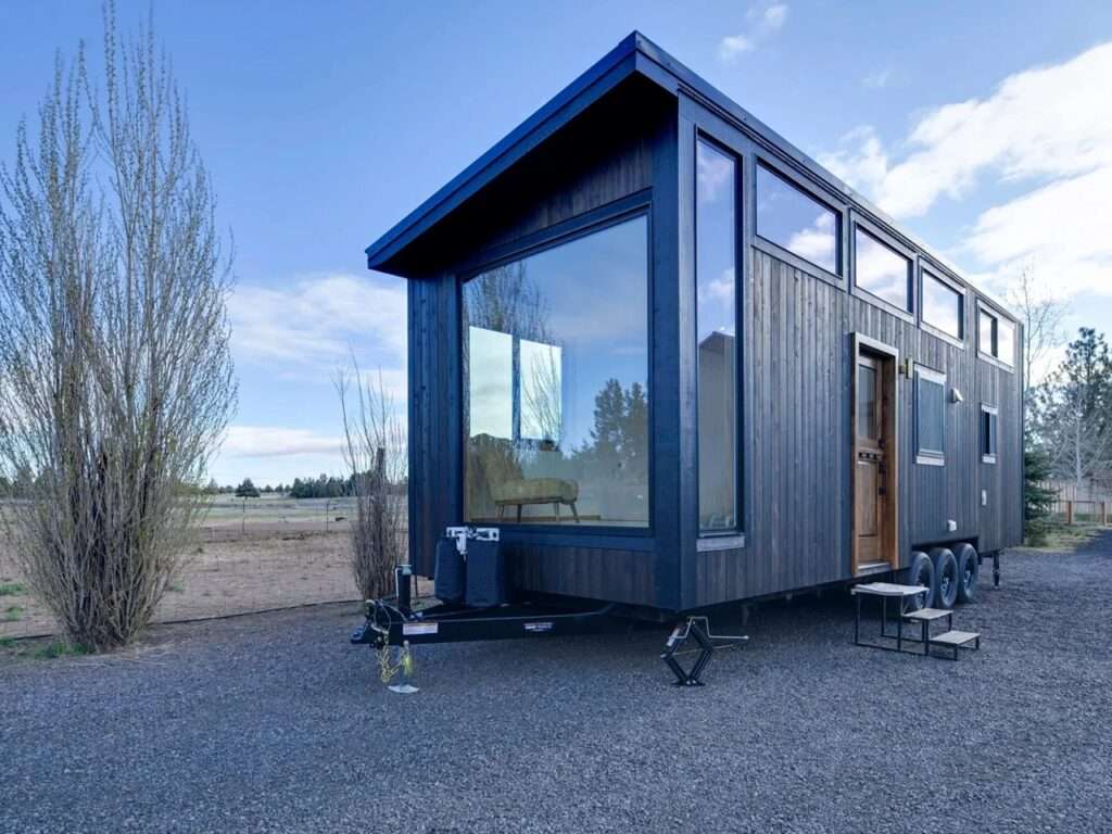 Exterior of Black Butte tiny home by Spindrift Homes featuring dark vertical wood siding, a triple-axle trailer, and large panoramic windows on a gravel plot.