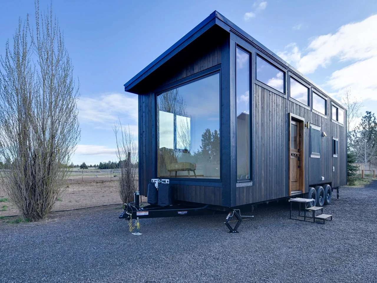 Exterior of Black Butte tiny home by Spindrift Homes featuring dark vertical wood siding, a triple-axle trailer, and large panoramic windows on a gravel plot.