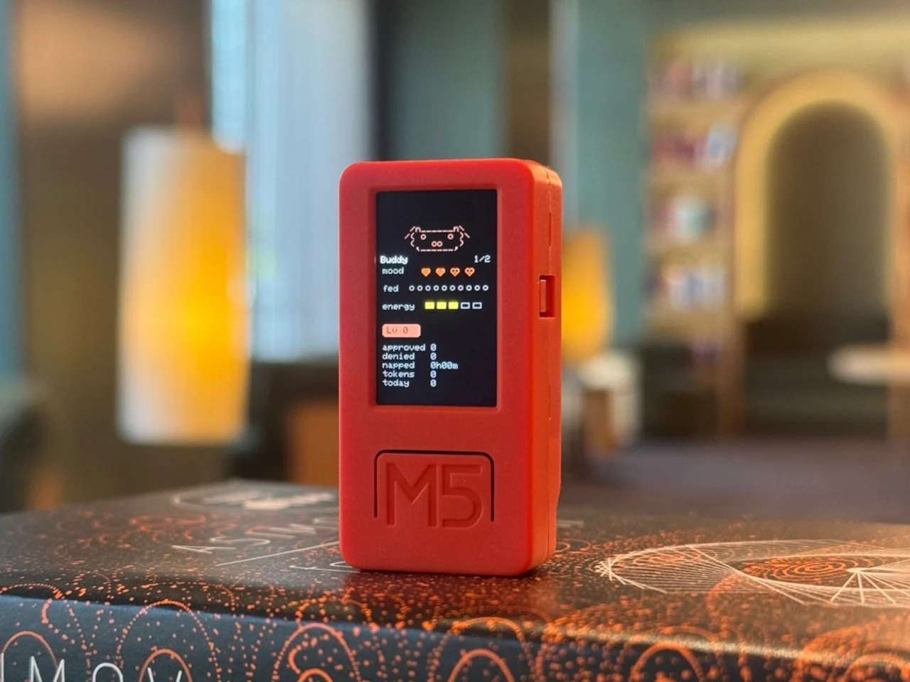 A close-up shot of the Claude Desktop Buddy hardware device in a bright orange case, standing vertically on a dark surface. The small color screen displays pixelated character states, stats like energy levels, and a digital interface.