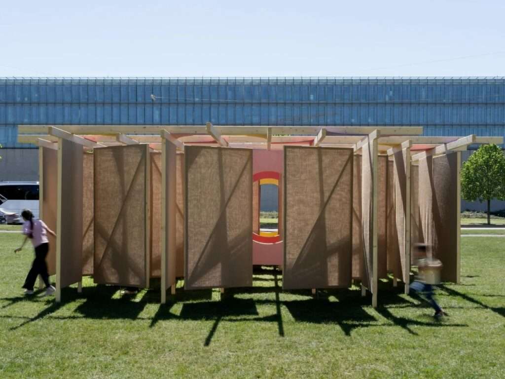 Ground-level view of the DELULU pavilion installation by Studio Carraldo on a green lawn, showing vertical jute fabric panels hanging from a light timber frame with children playing around it.