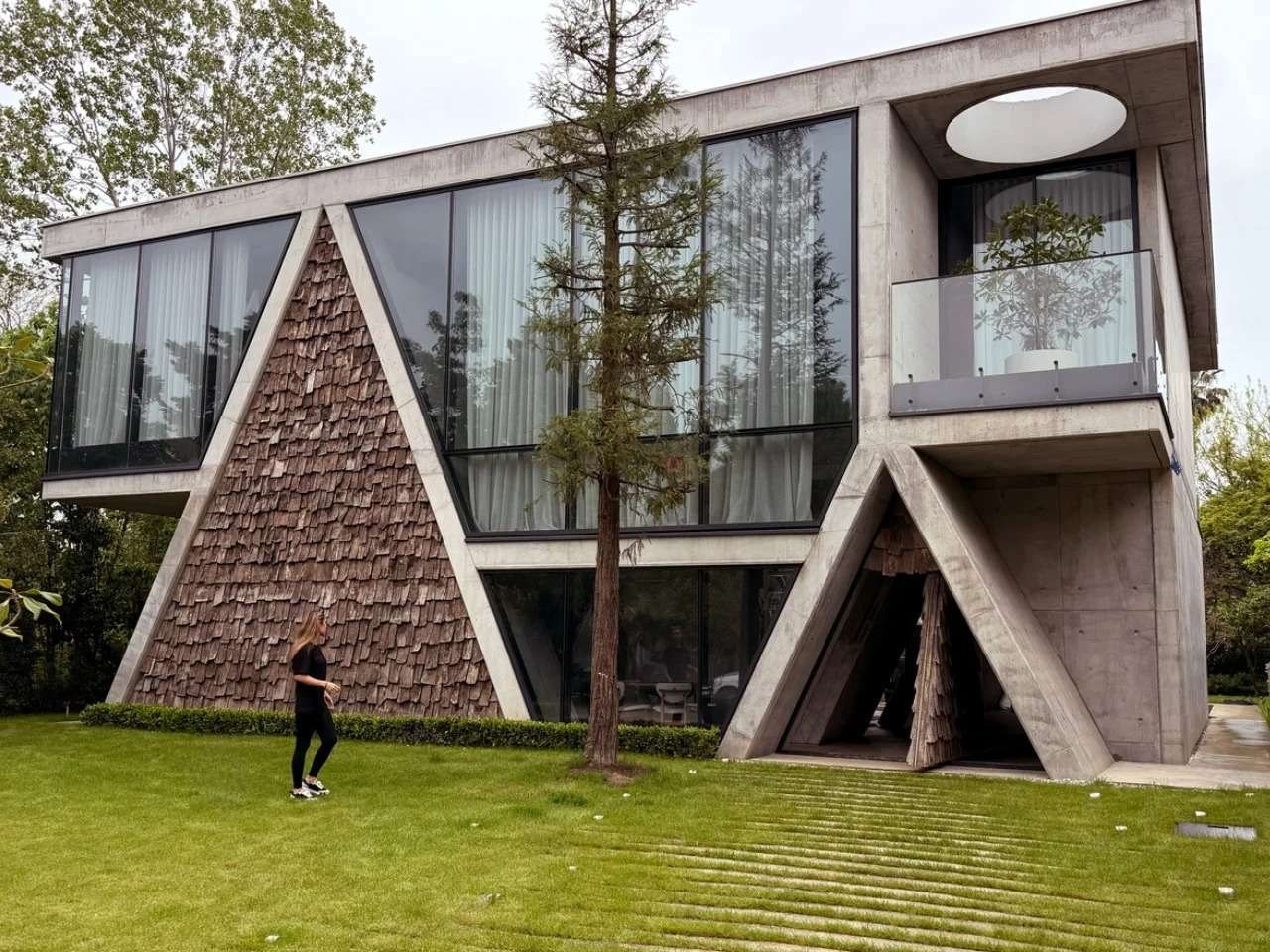 Modern concrete residential villa with a distinctive triangular wood-shingle accent wall and large floor-to-ceiling glass windows on a green lawn.