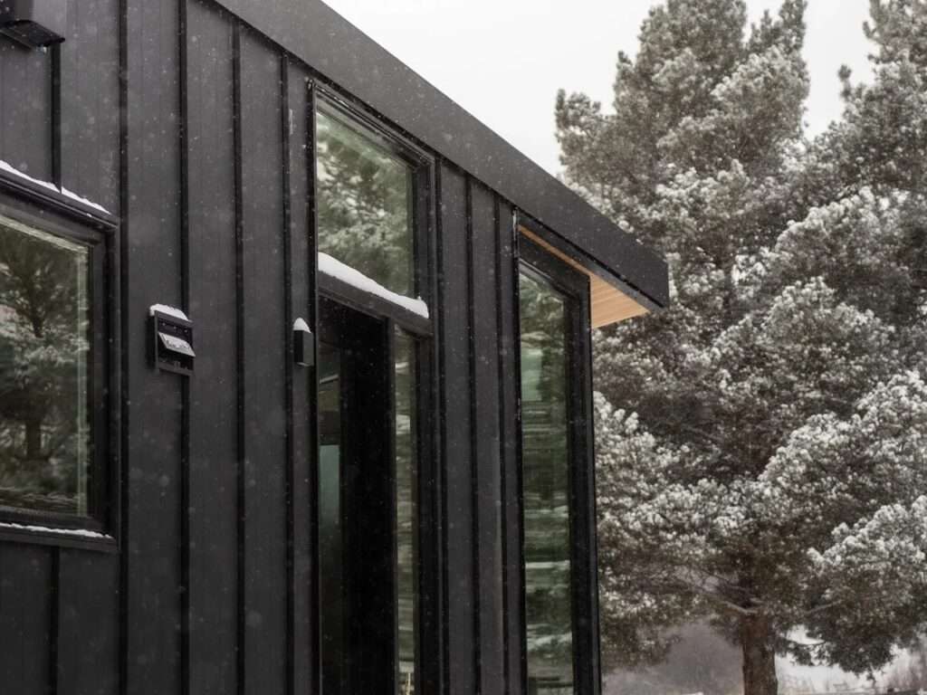 Close-up of the Halcyon Grand tiny home's exterior showing dark vertical metal cladding, glass windows, and a wood-paneled soffit under falling snow.
