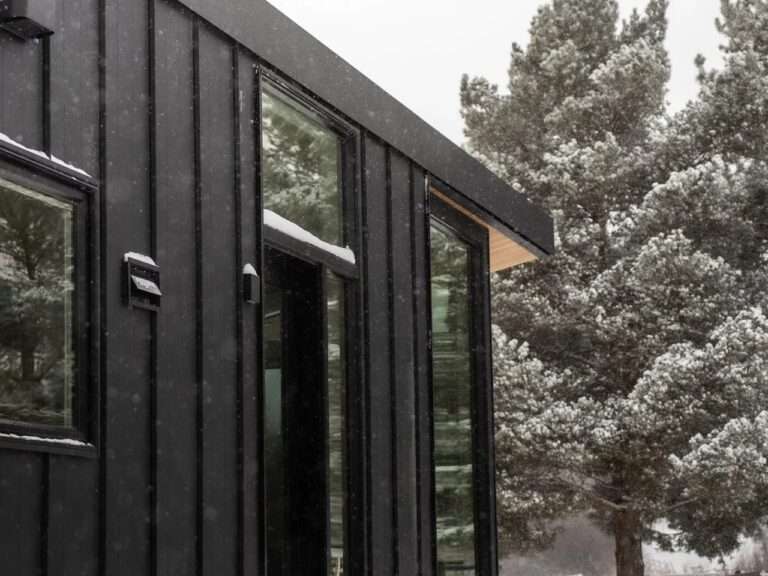 Close-up of the Halcyon Grand tiny home's exterior showing dark vertical metal cladding, glass windows, and a wood-paneled soffit under falling snow.