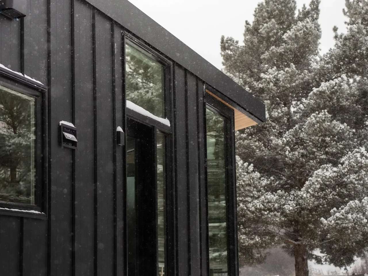Close-up of the Halcyon Grand tiny home's exterior showing dark vertical metal cladding, glass windows, and a wood-paneled soffit under falling snow.