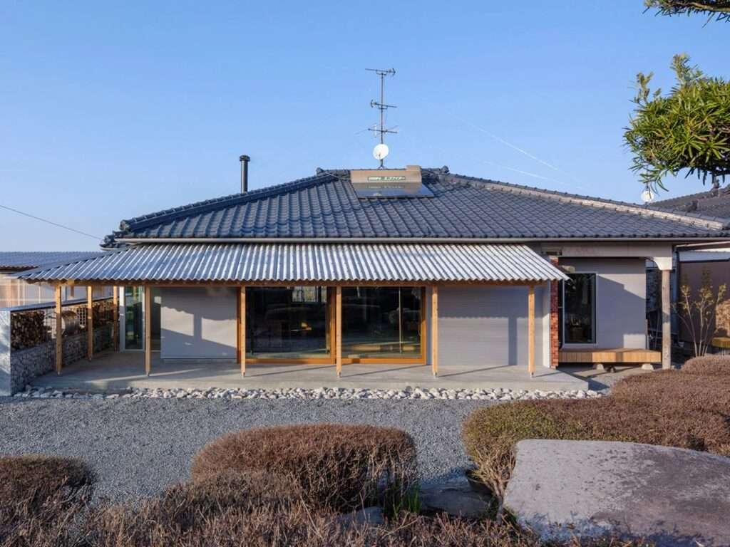 Traditional Japanese wooden house facade with a modern corrugated metal canopy extension, gravel courtyard, and large sliding glass doors under a dark tiled roof.