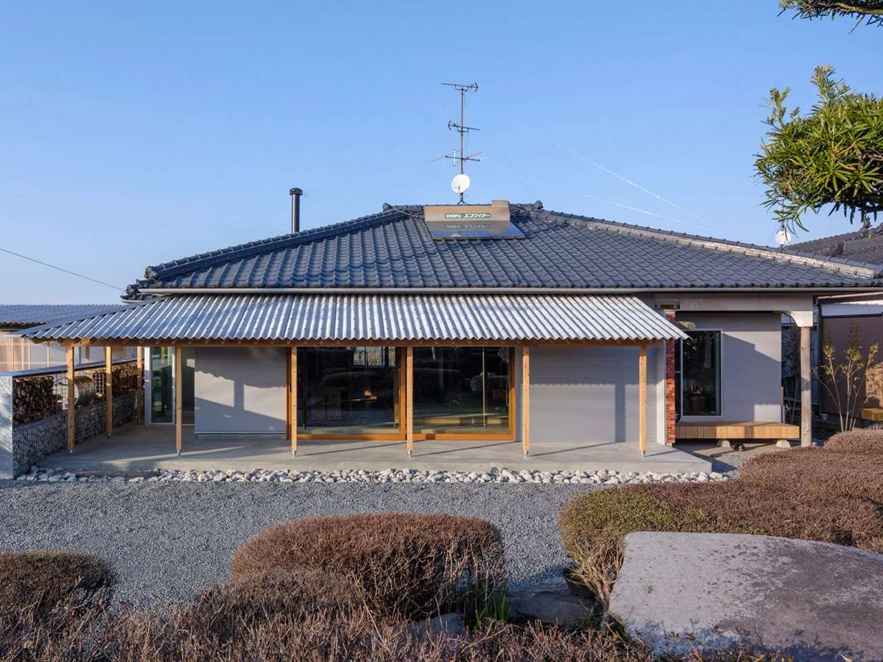 Traditional Japanese wooden house facade with a modern corrugated metal canopy extension, gravel courtyard, and large sliding glass doors under a dark tiled roof.