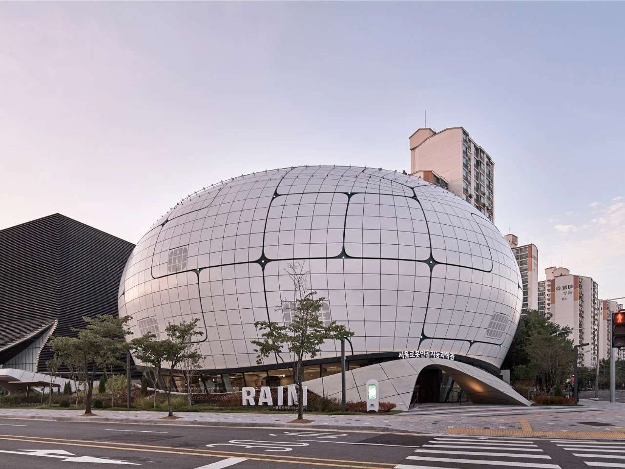 Daytime exterior view of the Seoul Robot and AI Museum (RAIM) featuring its spherical parametric envelope covered with grid-aligned curved metallic panels under a clear sky.