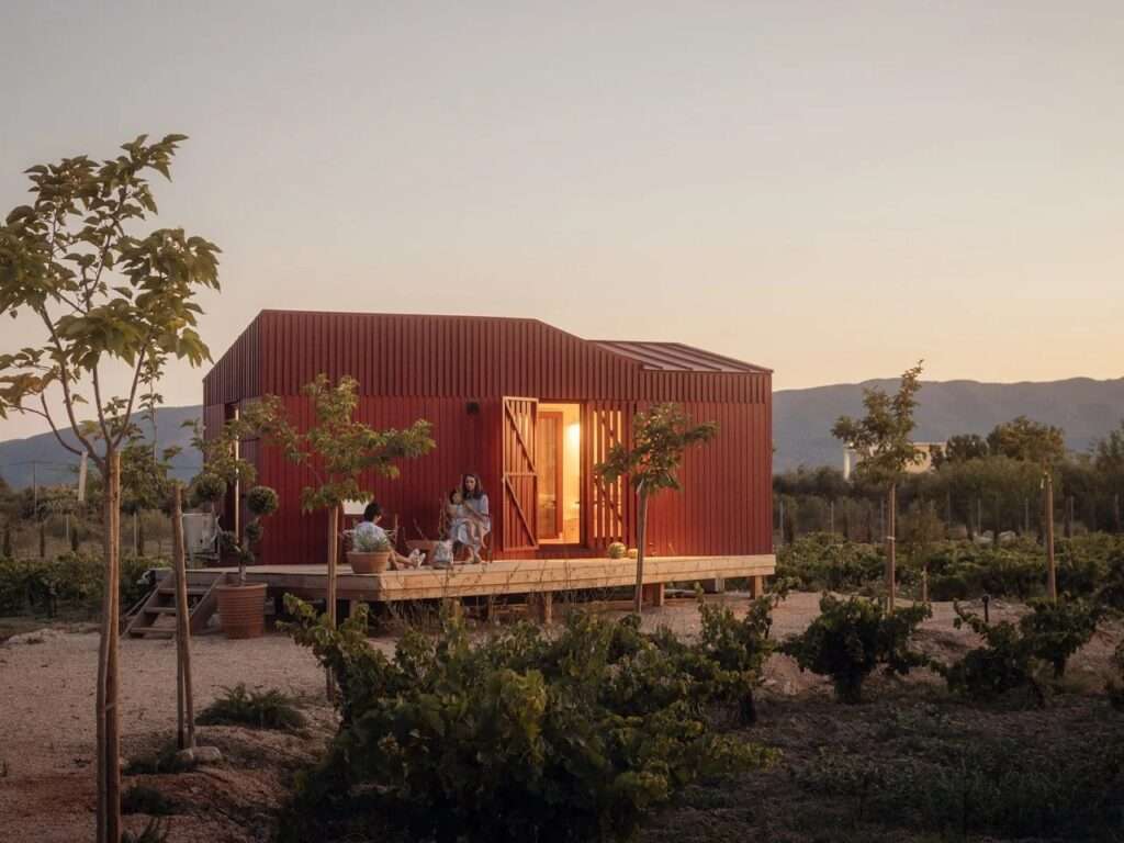Prefab Root Cabin by Kasawoo at sunset, featuring an illuminated interior through an open sliding glass door with a family sitting on the wooden patio deck.