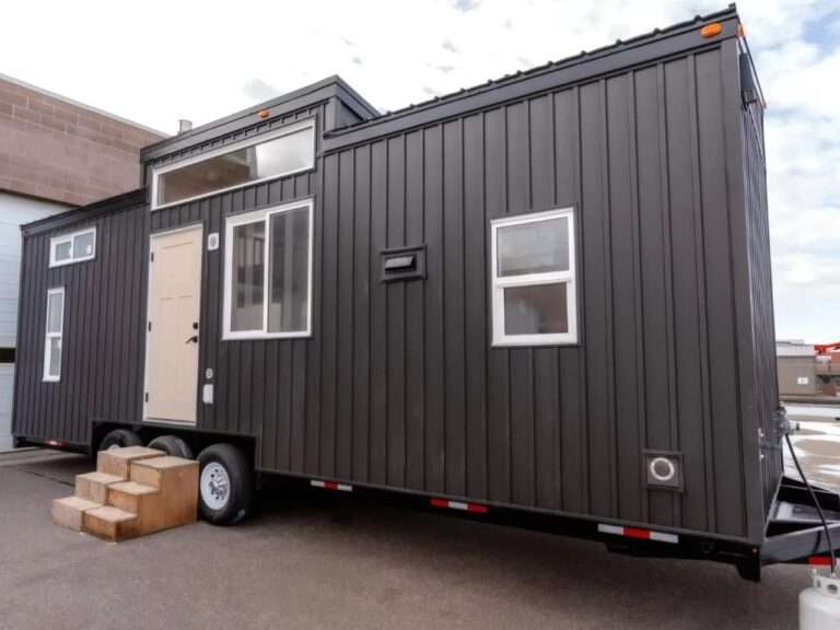 Exterior view of the Ruby tiny home by Teacup Tiny Homes featuring dark gray vertical metal siding, multiple windows, a light-colored entry door, and a triple-axle trailer chassis with wooden entry steps.