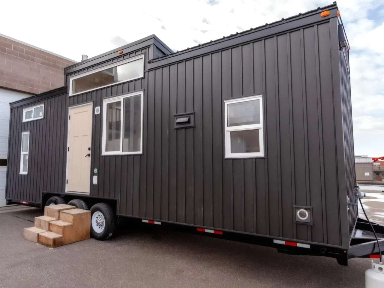 Exterior view of the Ruby tiny home by Teacup Tiny Homes featuring dark gray vertical metal siding, multiple windows, a light-colored entry door, and a triple-axle trailer chassis with wooden entry steps.