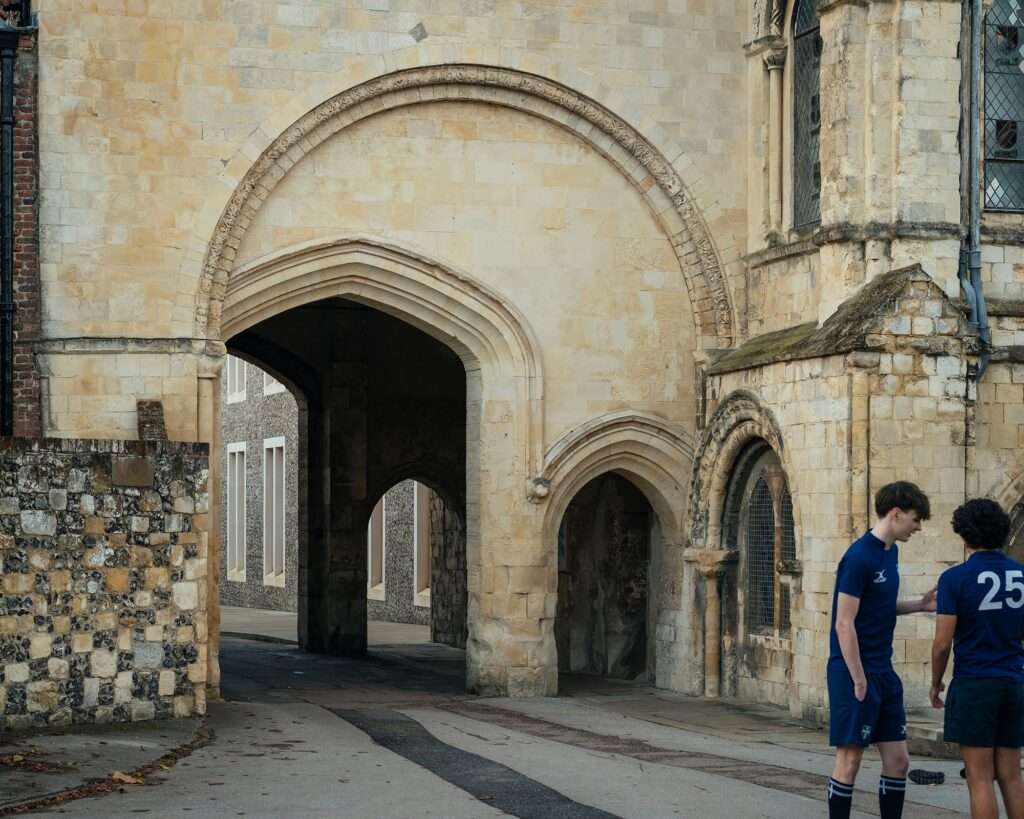 A historic stone archway gate with medieval architecture leading towards a glimpse of the modern flint stone facade of the Rausing Science Centre.