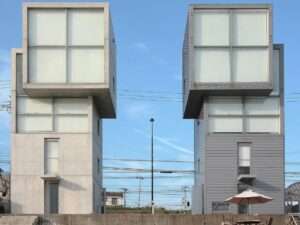 Front elevation of Tadao Ando's twin 4x4 Houses in Kobe, showing the original concrete tower on the left and the replicated wooden clad tower on the right against a clear blue sky.