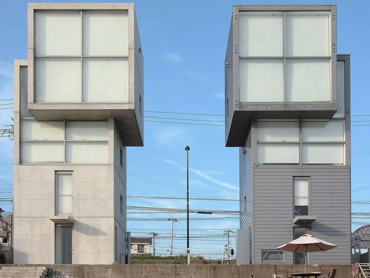 Front elevation of Tadao Ando's twin 4x4 Houses in Kobe, showing the original concrete tower on the left and the replicated wooden clad tower on the right against a clear blue sky.