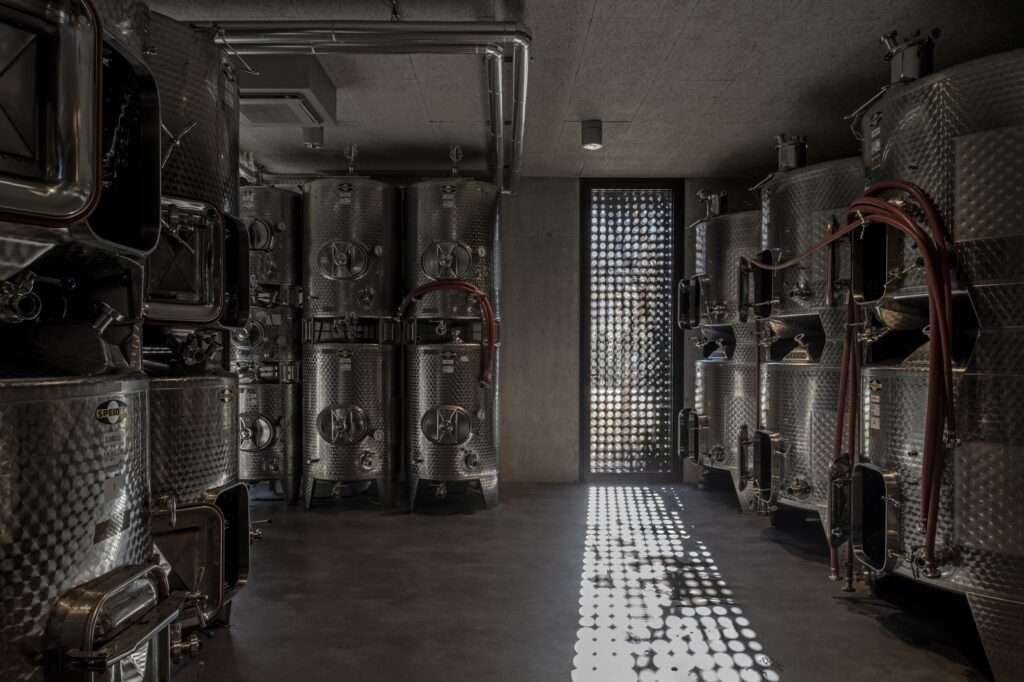 Industrial interior of Cantina Hauser showcasing stainless steel wine production tanks under light patterns filtering through the perforated facade.