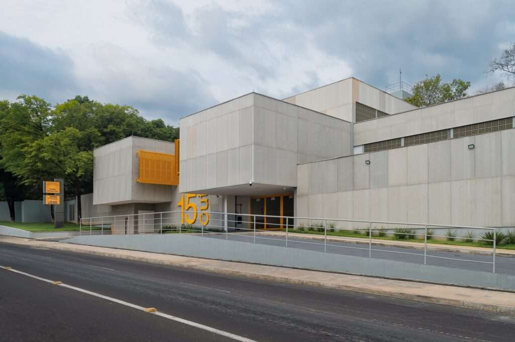 Wide angle street view of the Escola Municipal Ambiental 15 de Outubro in Teresina, showcasing the stepped concrete volume, access ramp, and surrounding green park trees.