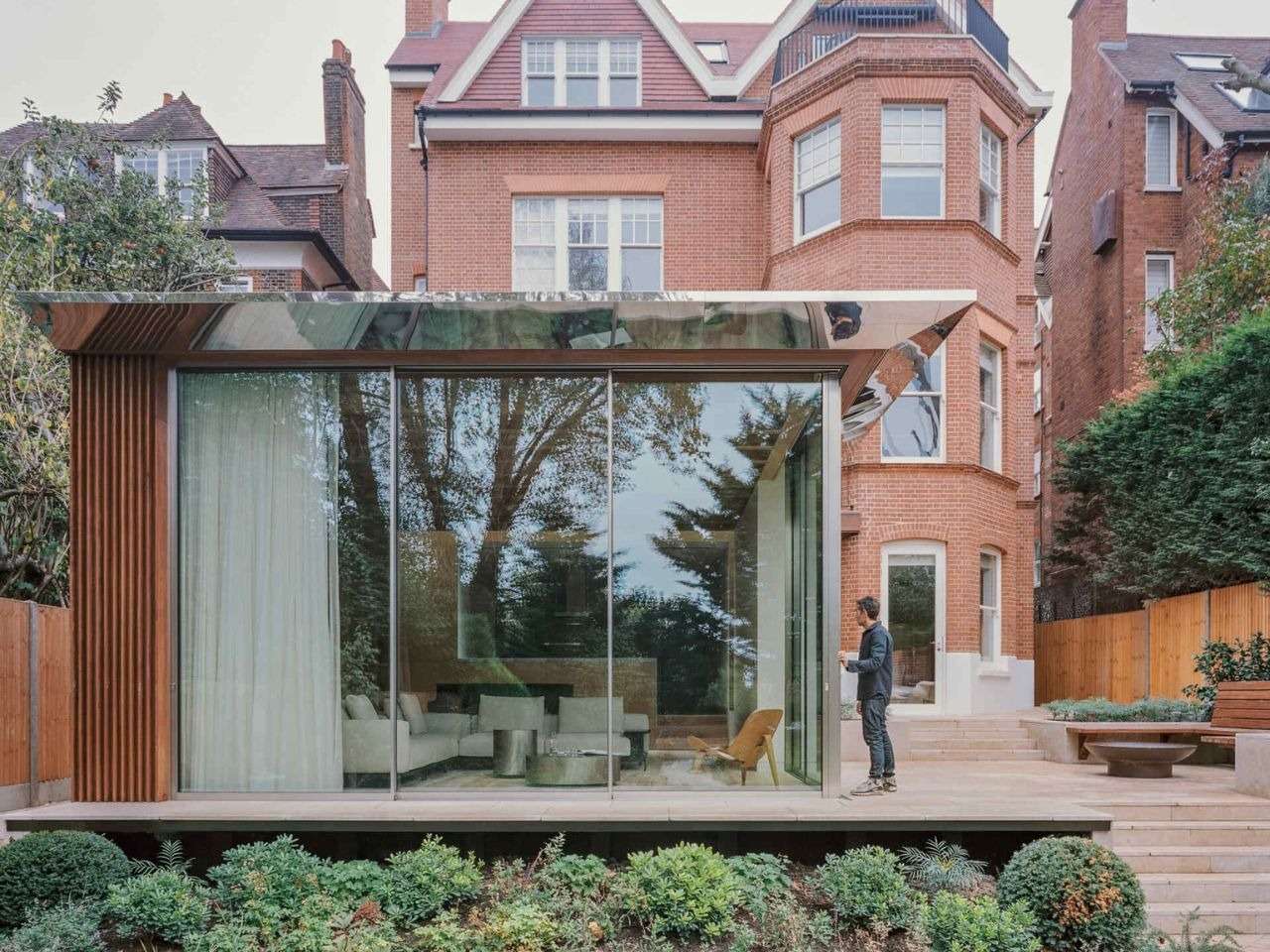 Modern glass extension with polished steel roof and Iroko wood cladding attached to a traditional brick house in Hampstead.