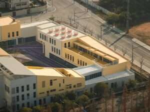 High-angle aerial view of the modular block kindergarten in Kunshan, showcasing stacked yellow, beige, and white educational volumes with a central courtyard and rooftop cylindrical light wells.