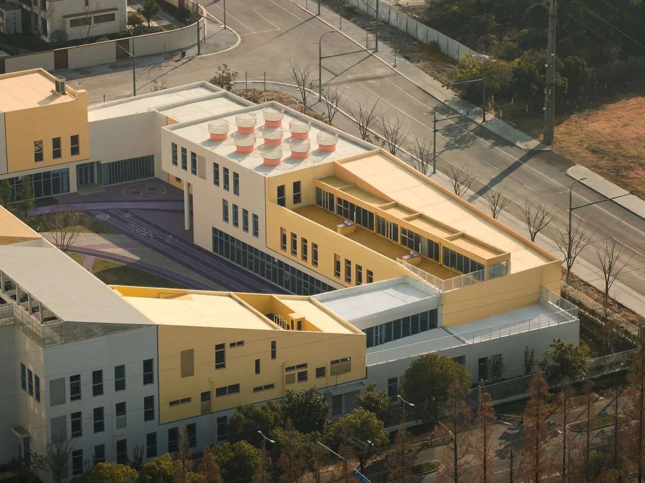 High-angle aerial view of the modular block kindergarten in Kunshan, showcasing stacked yellow, beige, and white educational volumes with a central courtyard and rooftop cylindrical light wells.