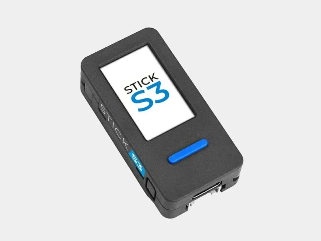 Studio product shot of a black M5StickS3 or Stick S3 hardware development kit device with a prominent blue physical button and a clear white screen displaying the text STICK S3.