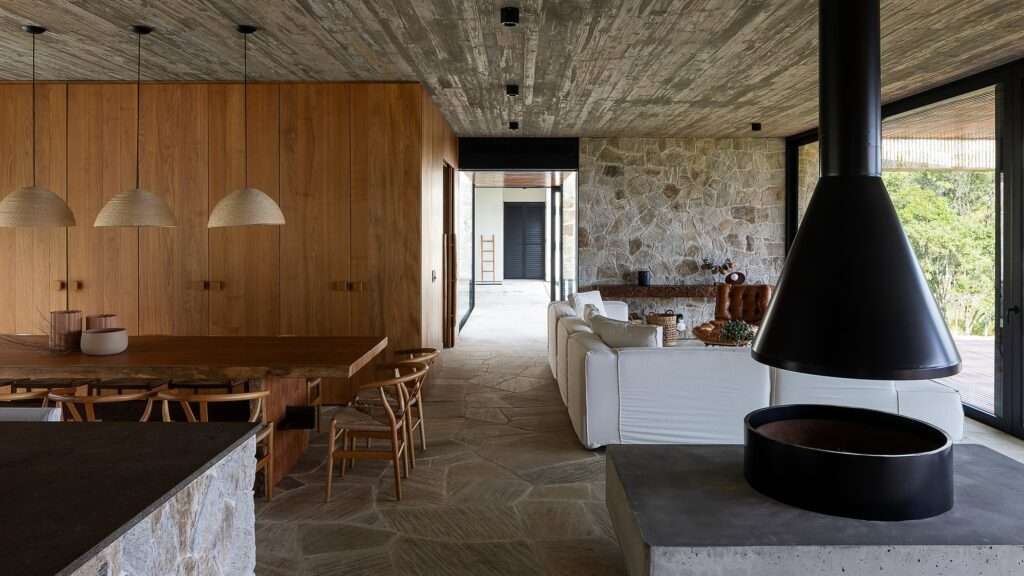 Dining room with long wooden table, woven pendant lights, and stone accent walls looking towards a central fireplace.