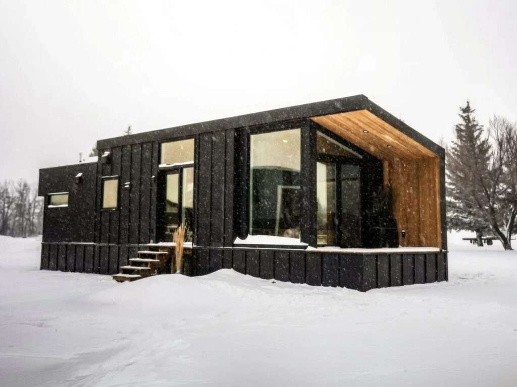 Full exterior view of the Halcyon Grand Park Model RV by Fritz Tiny Homes on a snow-covered landscape, featuring large glass facades and an integrated covered deck.