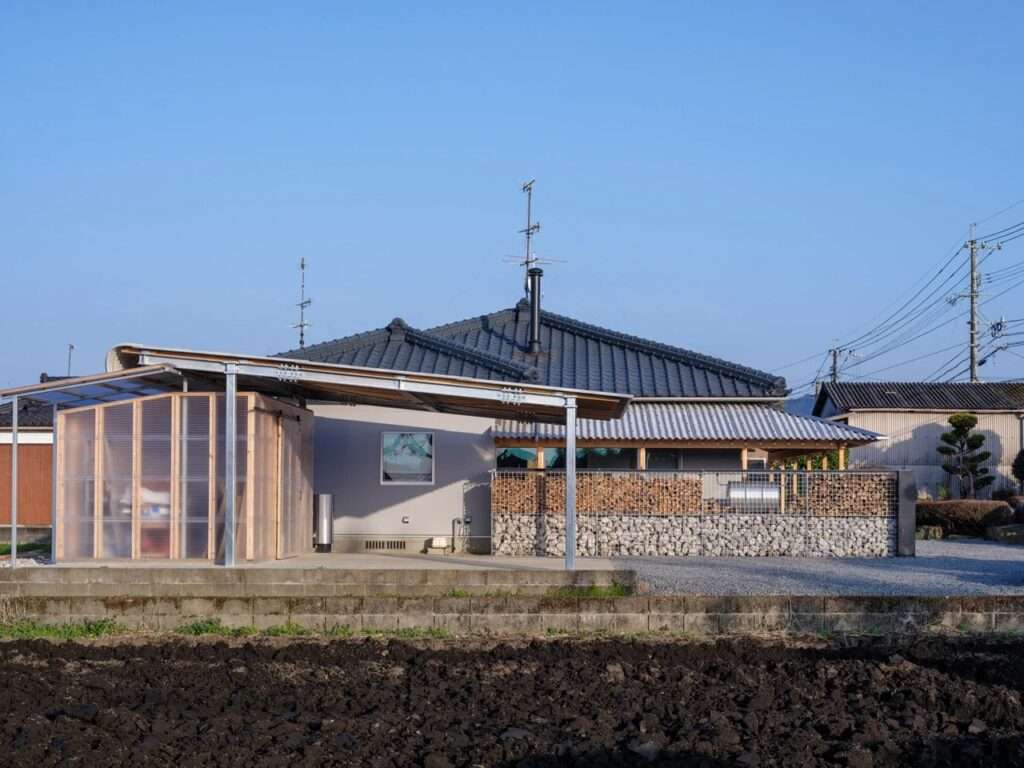 Side view of a renovated Japanese house featuring a stone gabion wall topped with stacked firewood, a translucent shelter, and an open gravel yard.