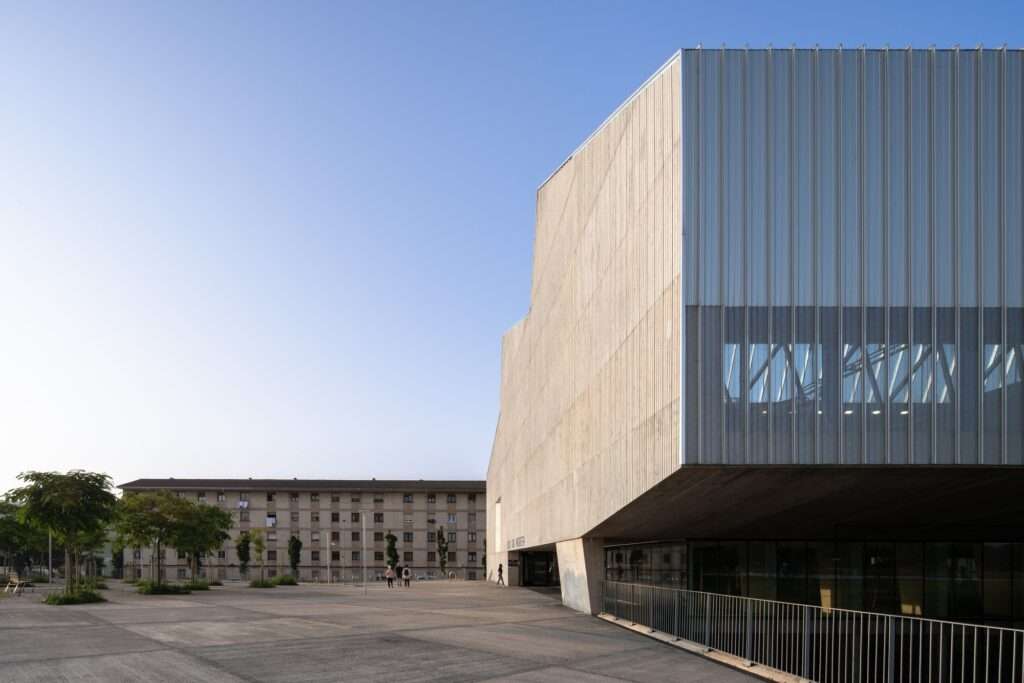Low-angle architectural view of the southern facade showing a massive board-formed concrete cantilever supporting a section clad in vertical perforated metal panels.