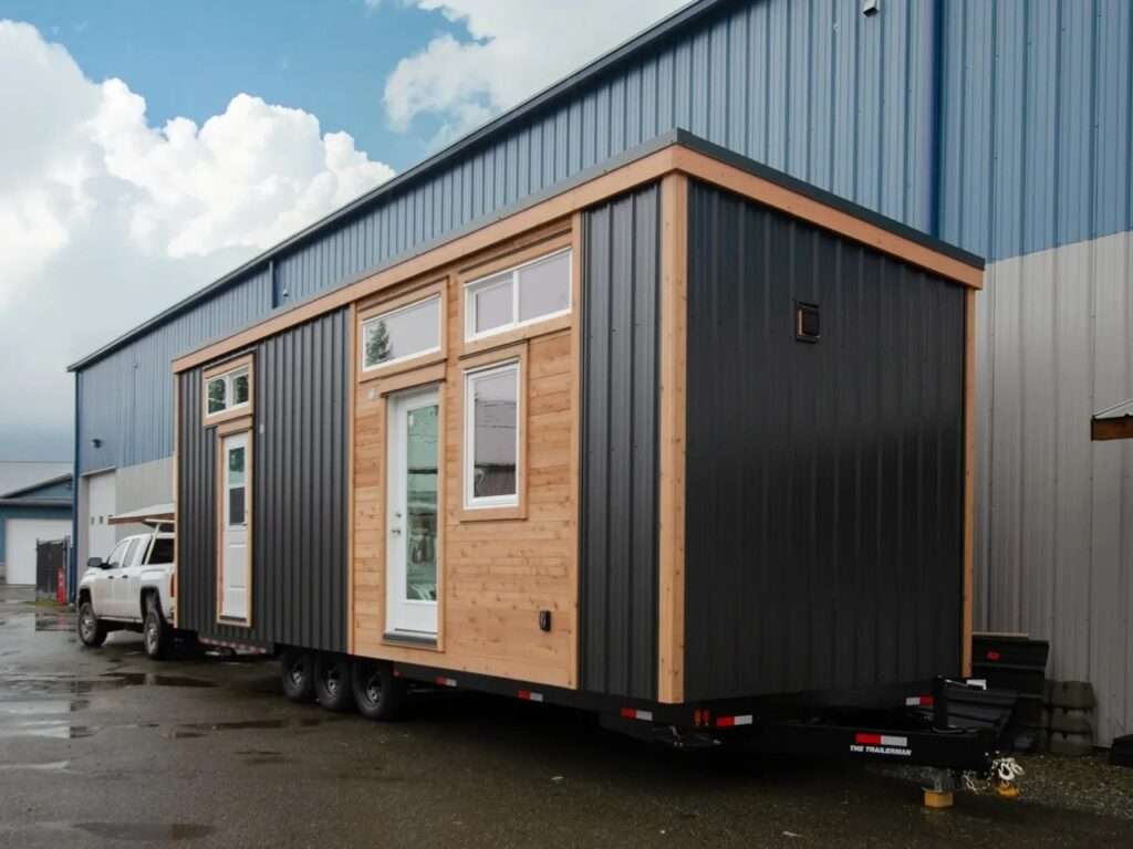 Exterior view of The Rose tiny home on a trailer, showcasing its modern West Coast facade combining dark metal cladding, warm cedar wood panels, and white-framed windows.