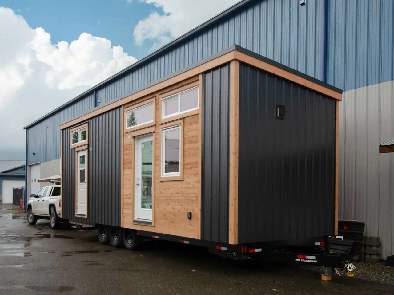 Exterior view of The Rose tiny home on a trailer, showcasing its modern West Coast facade combining dark metal cladding, warm cedar wood panels, and white-framed windows.
