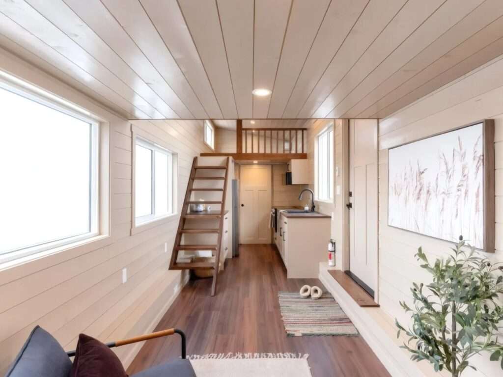Interior panoramic view of the Ruby tiny home displaying light wood shiplap walls and ceiling, hardwood floors, a kitchen counter, and a wooden ladder leading to a loft bedroom.