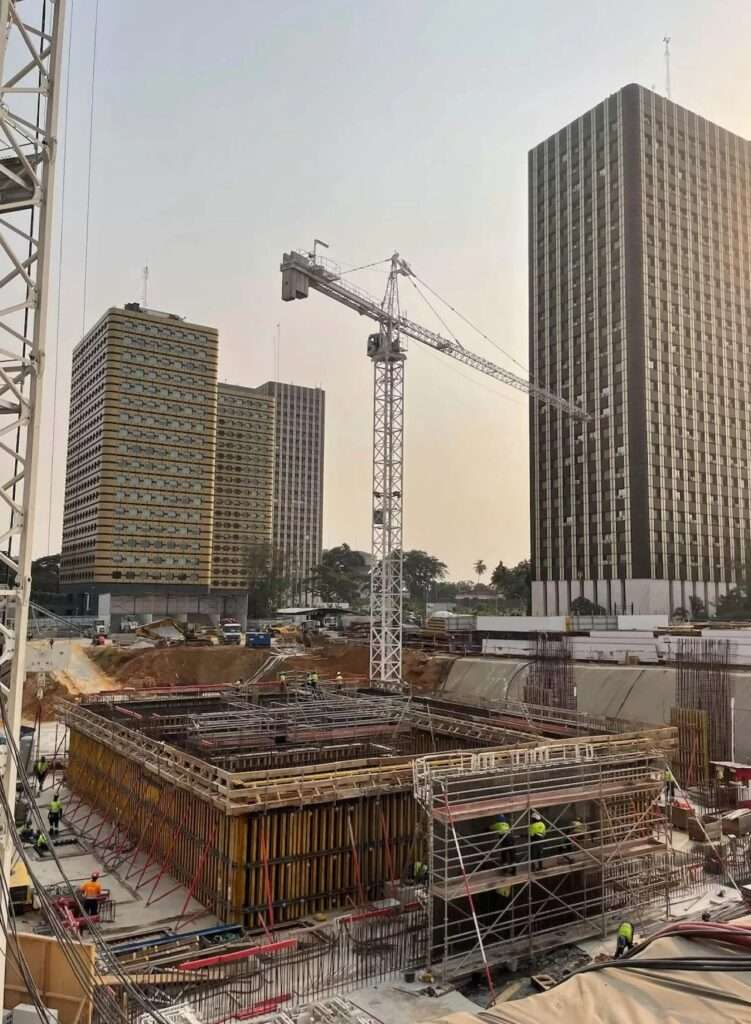 Construction site of Tour F Tower in Abidjan during foundation works, featuring a central crane, reinforced concrete formwork, and deep diaphragm wall installations with existing towers in the background.