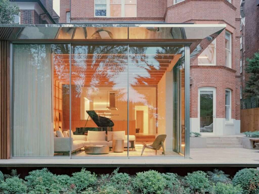 Dusk view of a modern glass-enclosed living room extension featuring integrated timber bookshelves, a grand piano, and warm internal lighting.